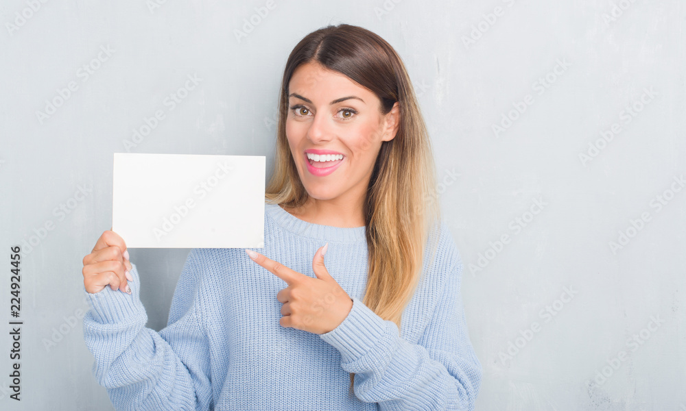 Young adult woman over grey grunge wall holding blank card very happy pointing with hand and finger Young adult woman over grey grunge wall holding blank card very happy pointing with hand and finger