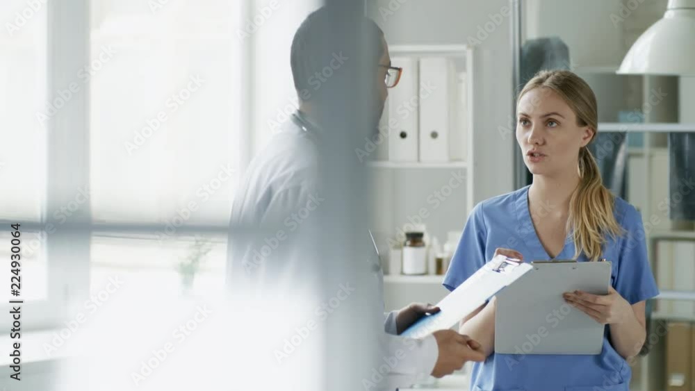 Tracking medium shot of two young doctors or scientists of different ethnicities holding clipboards with documents and talking to each other at work Tracking medium shot of two young doctors or scientists of different ethnicities holding clipboards with documents and talking to each other at work