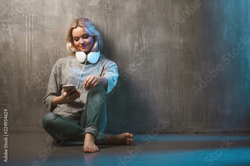 Young and attractive woman with smartphone and large headphones, sitting on the floor. Music in the mobile app.