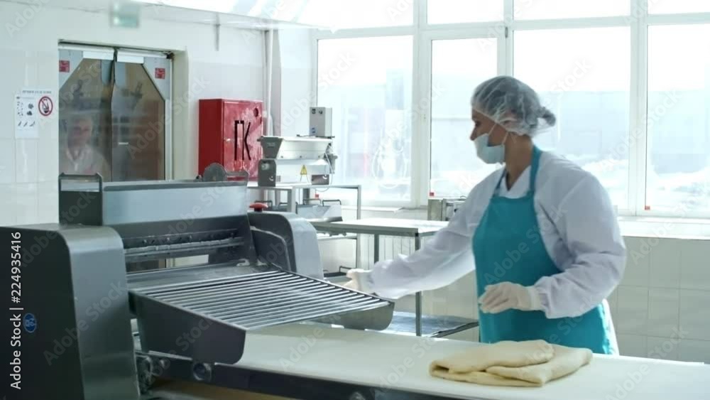 Woman in workwear, apron, bouffant mob cap, gloves and face mask operating dough sheeter when making laminated dough for croissants at factory Woman in workwear, apron, bouffant mob cap, gloves and face mask operating dough sheeter when making laminated dough for croissants at factory
