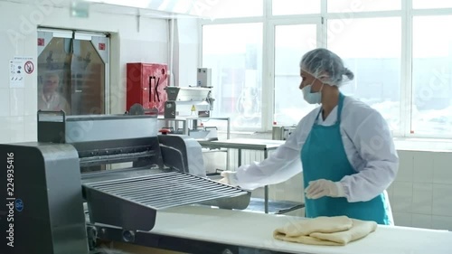 Woman in workwear, apron, bouffant mob cap, gloves and face mask operating dough sheeter when making laminated dough for croissants at factory photo