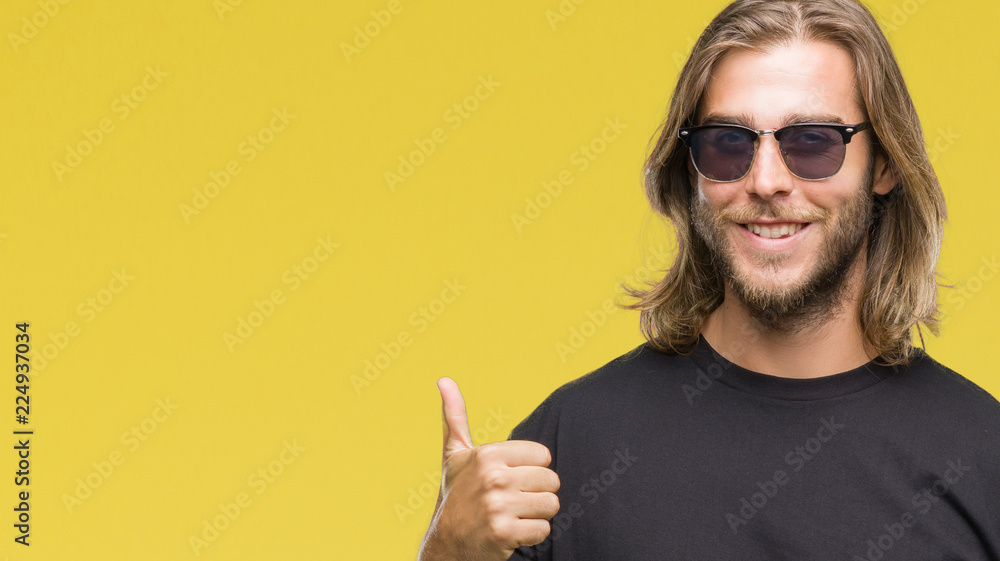 Young handsome man with long hair wearing sunglasses over isolated background doing happy thumbs up gesture with hand. Approving expression looking at the camera with showing success.
