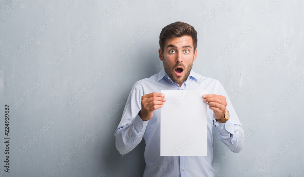 Handsome young man over grey grunge wall holding blank paper sheet contract scared in shock with a surprise face, afraid and excited with fear expression Handsome young man over grey grunge wall holding blank paper sheet contract scared in shock with a surprise face, afraid and excited with fear expression