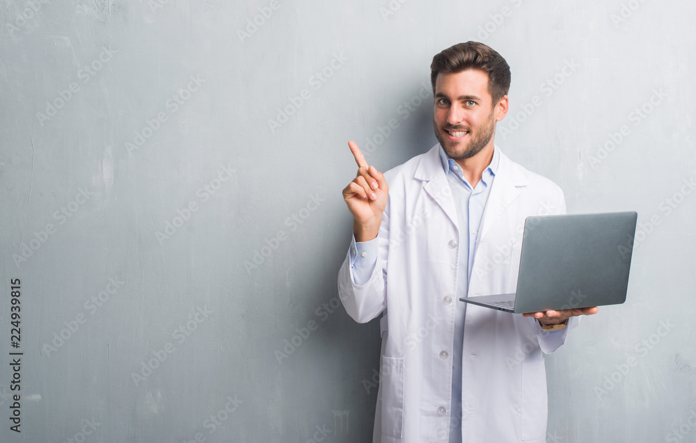 Handsome young man over grey grunge wall wearing white coat using laptop very happy pointing with hand and finger to the side Handsome young man over grey grunge wall wearing white coat using laptop very happy pointing with hand and finger to the side