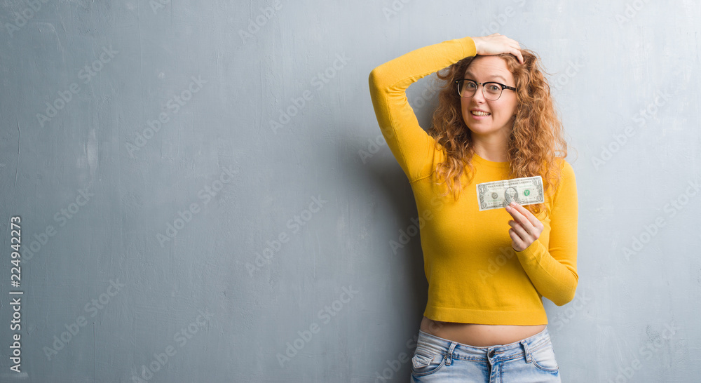 Young redhead woman over grey grunge wall holding a dollar stressed with hand on head, shocked with shame and surprise face, angry and frustrated. Fear and upset for mistake. Young redhead woman over grey grunge wall holding a dollar stressed with hand on head, shocked with shame and surprise face, angry and frustrated. Fear and upset for mistake.