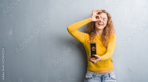 Young redhead woman over grey grunge wall holding gold ingot with happy face smiling doing ok sign with hand on eye looking through fingers