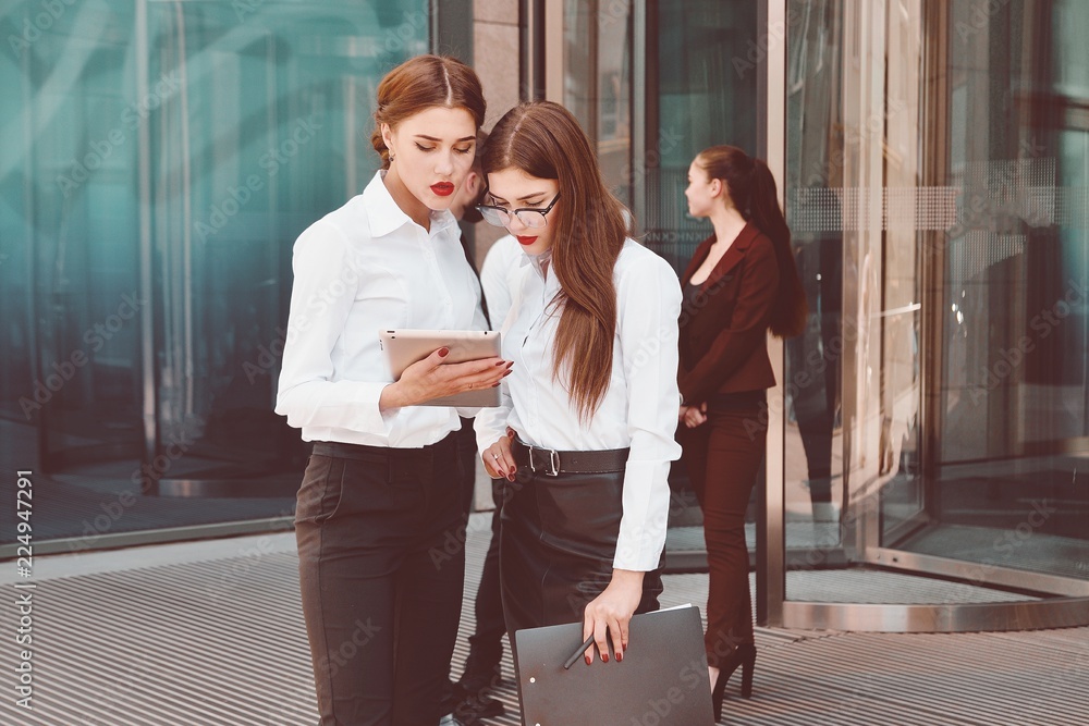 Business lady. Office staff. Two young girls with electronic tablets communicate against the background of a office building and a group of people. Electronic Gadget. Five people. 5.
