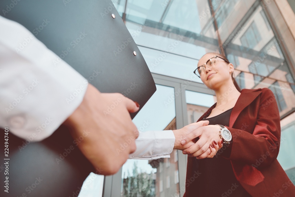 Obraz premium Handshake of two young girls against the background of a multi-storey office building. Make a deal. Friendly relations. Office staff