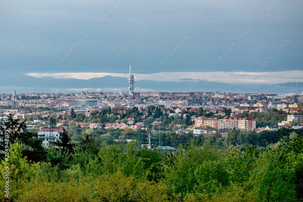 View of evening fall landscape of Czech republic capital city, Prague, Europe, famous tall observation tower at Zizkov, houses, horizon in distance, grey blue cloudy sky, green tree foreground