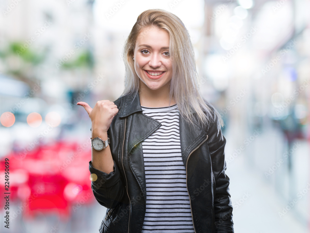 Young blonde woman wearing fashion jacket over isolated background smiling with happy face looking and pointing to the side with thumb up.