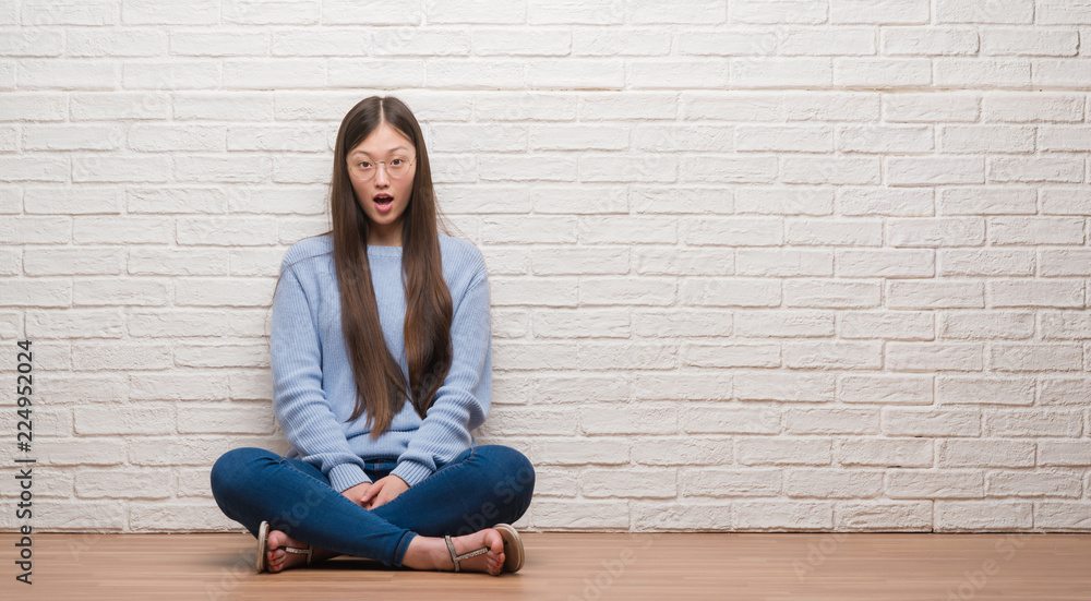 Young Chinese woman sitting on the floor over brick wall afraid and shocked with surprise expression, fear and excited face. Young Chinese woman sitting on the floor over brick wall afraid and shocked with surprise expression, fear and excited face.