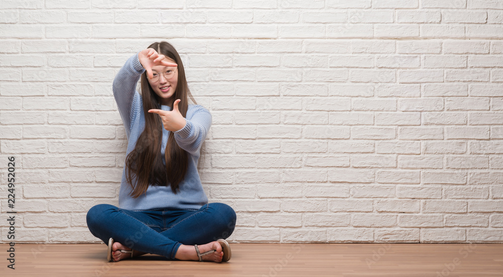 Young Chinese woman sitting on the floor over brick wall smiling making frame with hands and fingers with happy face. Creativity and photography concept. Young Chinese woman sitting on the floor over brick wall smiling making frame with hands and fingers with happy face. Creativity and photography concept.
