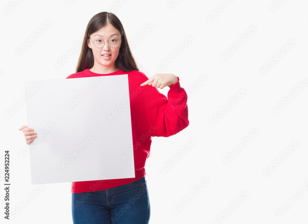 Young Chinese woman over isolated background holding blank banner with surprise face pointing finger to himself Young Chinese woman over isolated background holding blank banner with surprise face pointing finger to himself