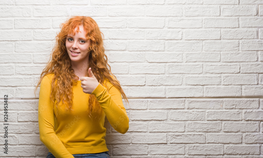 Young redhead woman standing over brick wall happy with big smile doing ok sign, thumb up with fingers, excellent sign Young redhead woman standing over brick wall happy with big smile doing ok sign, thumb up with fingers, excellent sign