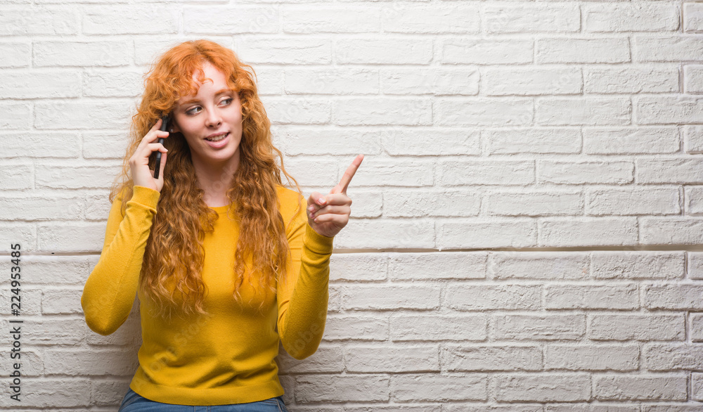 Young redhead woman standing over brick wall talking on the phone very happy pointing with hand and finger to the side Young redhead woman standing over brick wall talking on the phone very happy pointing with hand and finger to the side