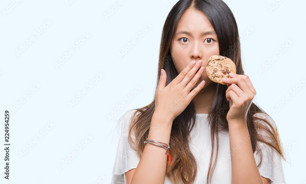 Young asian woman eating chocolate chip cookie over isolated background cover mouth with hand shocked with shame for mistake, expression of fear, scared in silence, secret concept Young asian woman eating chocolate chip cookie over isolated background cover mouth with hand shocked with shame for mistake, expression of fear, scared in silence, secret concept