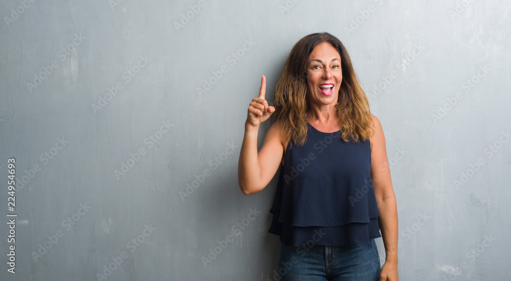 Middle age hispanic woman standing over grey grunge wall pointing finger up with successful idea. Exited and happy. Number one. Middle age hispanic woman standing over grey grunge wall pointing finger up with successful idea. Exited and happy. Number one.