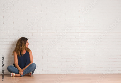 Middle age hispanic woman sitting on the floor over white brick wall looking to side, relax profile pose with natural face with confident smile. © Krakenimages.com