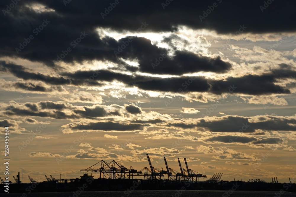 Dramatic clouds over the New Jersey waterfront at sunset, with silhouettes of machinery at the docks.