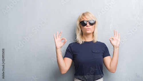 Adult caucasian woman over grunge grey wall wearing sunglasses relax and smiling with eyes closed doing meditation gesture with fingers. Yoga concept.