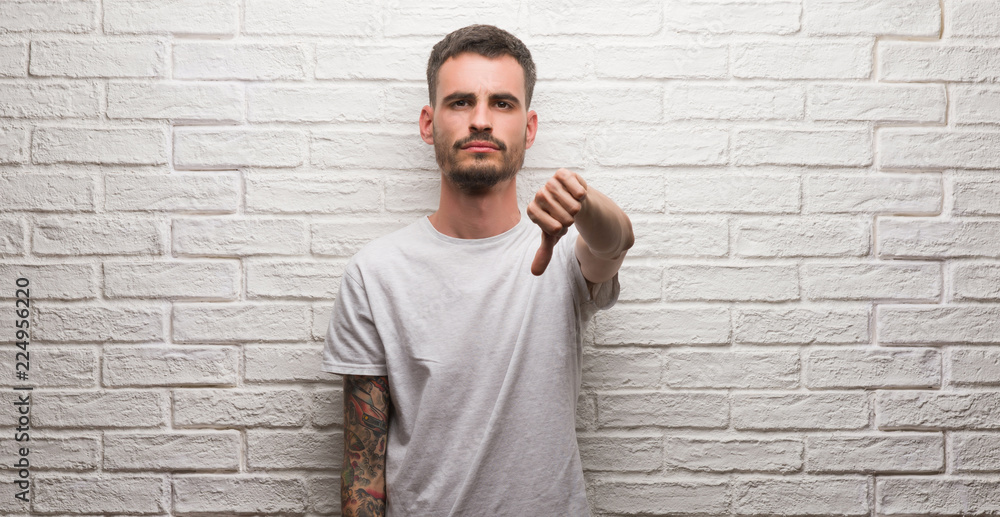 Young adult man standing over white brick wall with angry face, negative sign showing dislike with thumbs down, rejection concept Young adult man standing over white brick wall with angry face, negative sign showing dislike with thumbs down, rejection concept