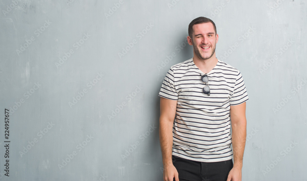 Young caucasian man over grey grunge wall winking looking at the camera with sexy expression, cheerful and happy face. Young caucasian man over grey grunge wall winking looking at the camera with sexy expression, cheerful and happy face.