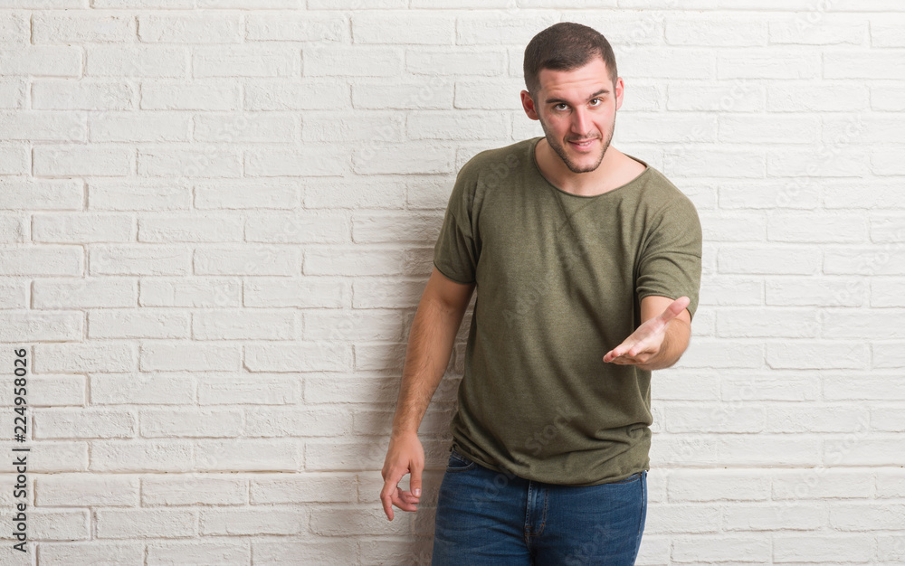 Young caucasian man standing over white brick wall smiling friendly offering handshake as greeting and welcoming. Successful business. Young caucasian man standing over white brick wall smiling friendly offering handshake as greeting and welcoming. Successful business.