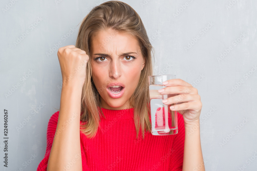 Beautiful young woman over grunge grey wall drinking a glass of water annoyed and frustrated shouting with anger, crazy and yelling with raised hand, anger concept Beautiful young woman over grunge grey wall drinking a glass of water annoyed and frustrated shouting with anger, crazy and yelling with raised hand, anger concept