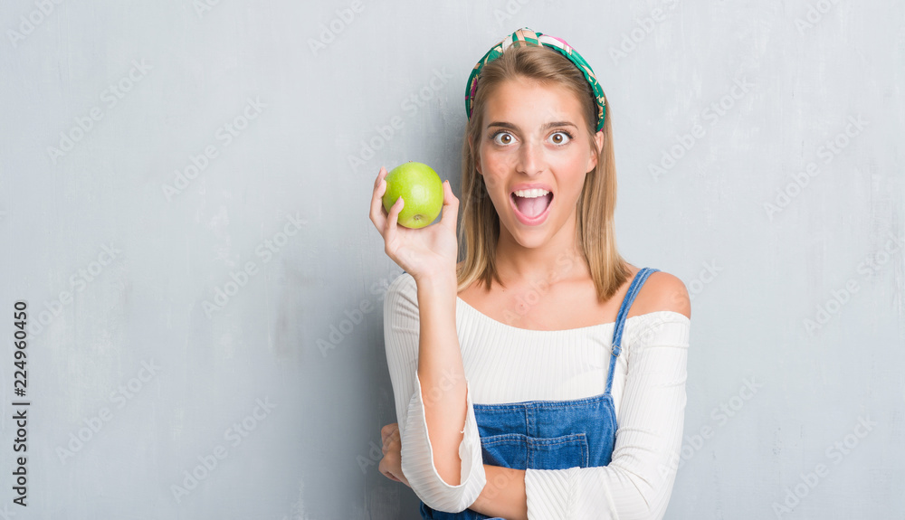 Beautiful young woman over grunge grey wall eating green apple scared in shock with a surprise face, afraid and excited with fear expression Beautiful young woman over grunge grey wall eating green apple scared in shock with a surprise face, afraid and excited with fear expression