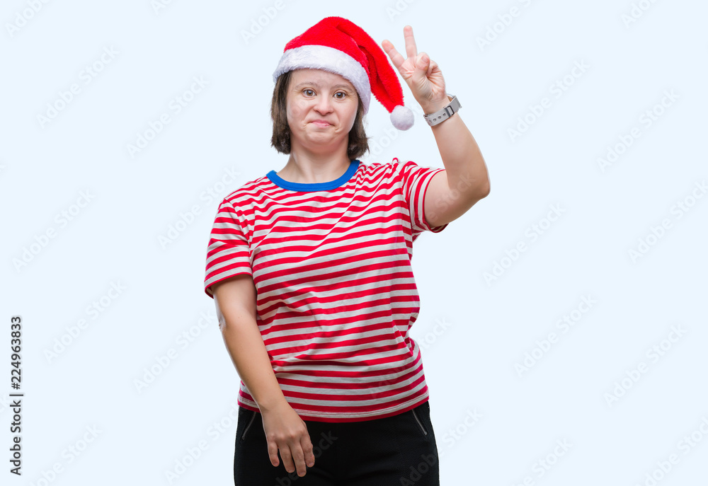 Young adult woman with down syndrome wearing christmas hat over isolated background smiling with happy face winking at the camera doing victory sign. Number two. Young adult woman with down syndrome wearing christmas hat over isolated background smiling with happy face winking at the camera doing victory sign. Number two.