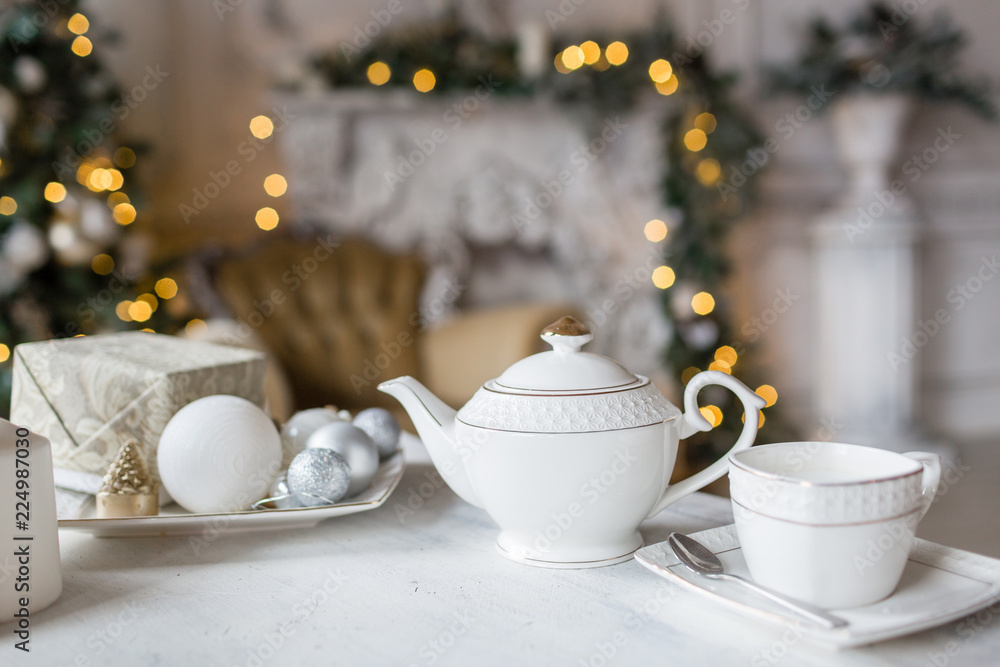 Fototapeta premium Tea time, white teapot and cups on the table. Decorated Christmas tree. Classic apartments with a white fireplace.