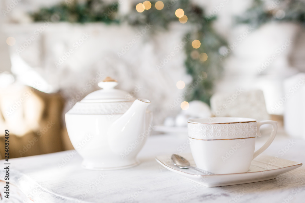 Tea time, white teapot and cups on the table. Decorated Christmas tree. Classic apartments with a white fireplace.