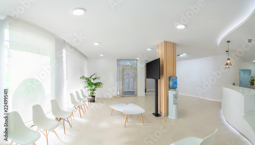 Panorama of a bright reception and waiting room in a clinic with desk, modern chairs and plants. Indoor mockup with screen with copy space. photo