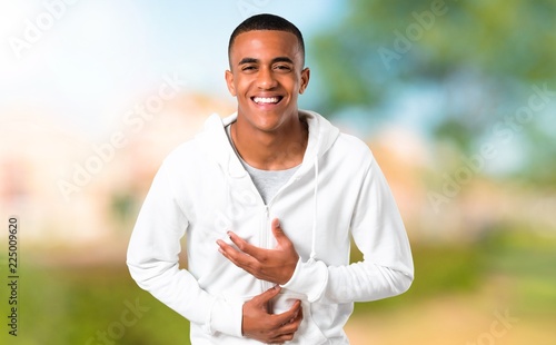 Dark-skinned young man with white sweatshirt smiling a lot while putting hands on chest in a park