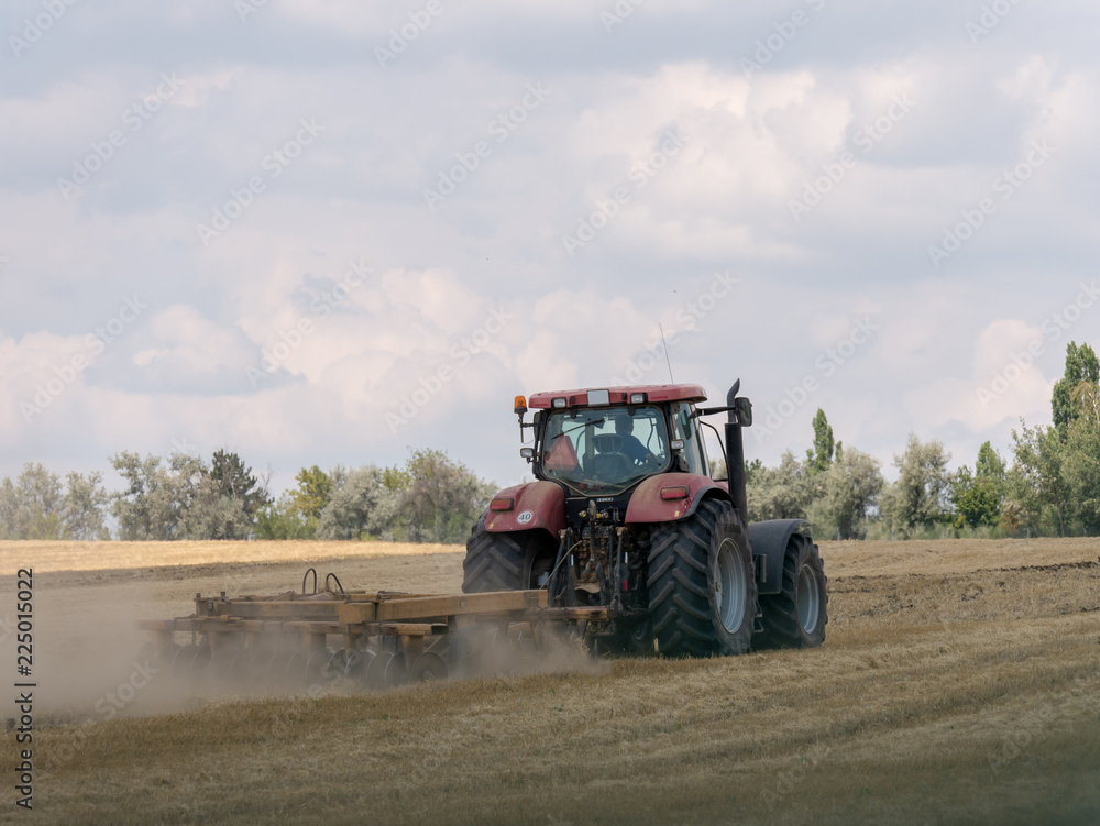 Fototapeta premium Red tractor with plow on harvest field. Summer field and red tractor. Tractor on harvest field. Lowing after harvest. Plowing on the field.