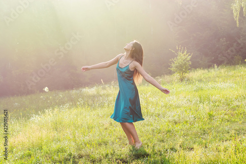 Beautiful smiling young woman is dancing and whirls barefoot on a flower meadow at sunset.
