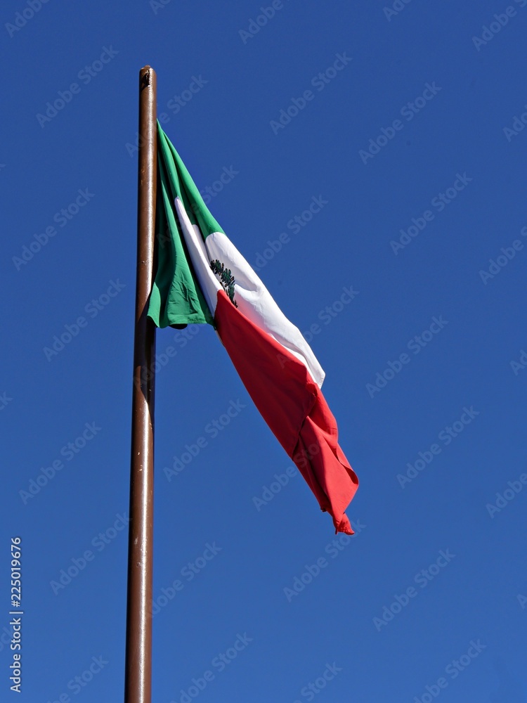 Flag of Mexico flying halfway unfurled in the air against a backdrop of blue skies