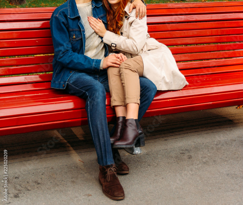 couple sitting on a bench, legs of a girl on a guy, a man hugging a woman.a guy in jeans and a denim jacket, a girl in a beige coat and pants, brown shoes