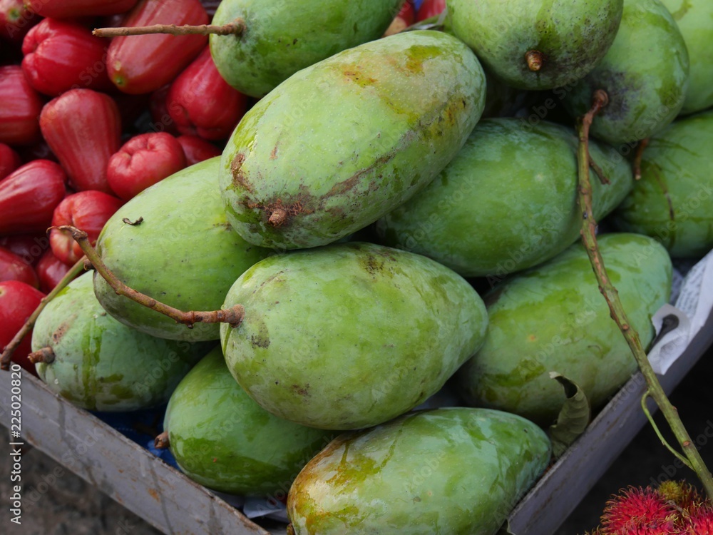 Close up of green unripe mangoes with rose apples in a wooden crate at an outdoor market Close up of green unripe mangoes with rose apples in a wooden crate at an outdoor market