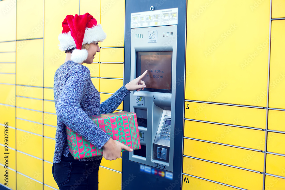 Woman with santa hat client using automated self service post terminal machine or locker to deposit the parcel for storage Woman with santa hat client using automated self service post terminal machine or locker to deposit the parcel for storage