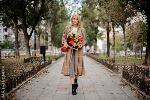 Beautiful blonde woman in plaid coat holding a bright bouquet of flowers standing on alley
