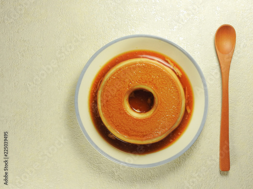 Top view of delicious handmade yellow color round egg pudding dessert with caramel sauce on blue side of the white plate beside the wooden brown spoon isolated on white background.Sweet food.