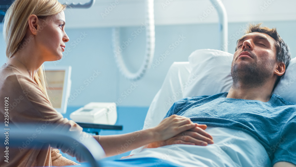 Fototapeta premium In the Hospital Sick Man Lying on the Bed, His Visiting Wife Hopefully Sits Beside Him Holds His Hands and Hopes for Recovery.