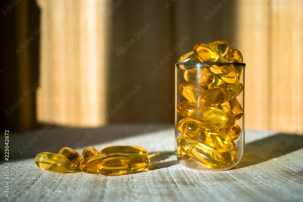 Yellow gold oil vitamin, omega 3 capsules in a glass beaker in the rays of light on a wooden background. Close up, selective focus Yellow gold oil vitamin, omega 3 capsules in a glass beaker in the rays of light on a wooden background. Close up, selective focus