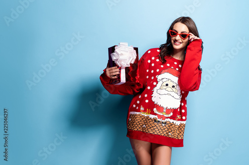 Pretty brunette woman in sunglasses and red oversize pullover with Santa design holding gift box while standing on blue background in studio. Isolate