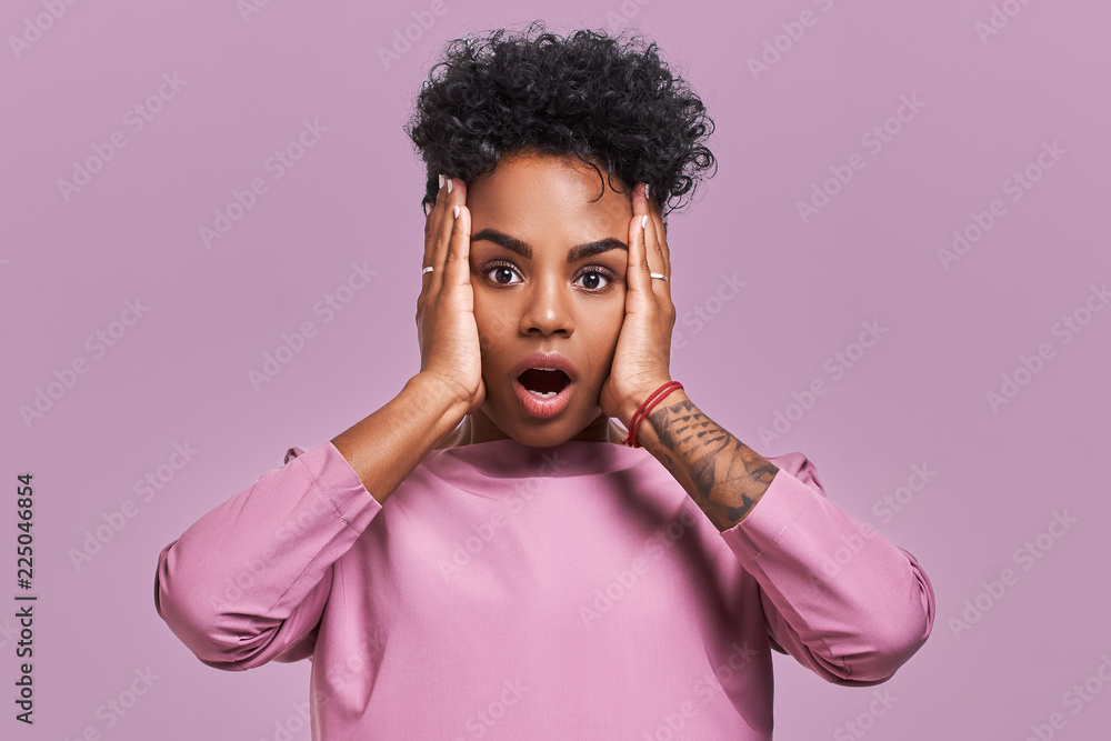 Young lovely dark skinned female has curly hair, looks surprisingly at camera, keeps jaw dropped, wonders sudden news, poses against lavender background. Facial expressions Young lovely dark skinned female has curly hair, looks surprisingly at camera, keeps jaw dropped, wonders sudden news, poses against lavender background. Facial expressions
