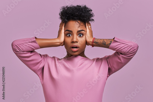 Stunned African American female stares at camera and keeps mouth opened, scratches head and looks surprisingly, realizes bad news, stands against lavender background. People, reaction, ethnicity