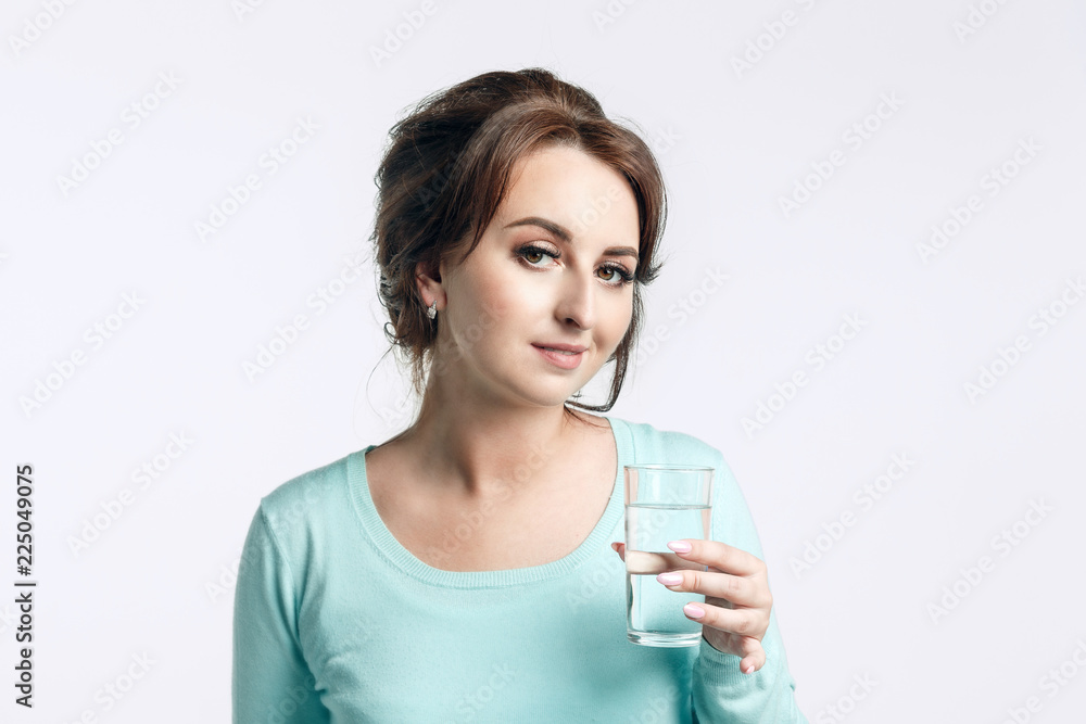 A young brown-haired woman in a mint-colored jumper holding a glass of clean water and looking into the camera with a slight smile. A young brown-haired woman in a mint-colored jumper holding a glass of clean water and looking into the camera with a slight smile.