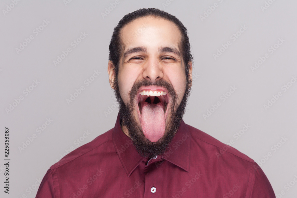 Portrait of funny crazy handsome man with beard in red shirt looking at camera with big open mouth and tongue out and screaming or shouting. indoor studio shot, isolated on gray background. Portrait of funny crazy handsome man with beard in red shirt looking at camera with big open mouth and tongue out and screaming or shouting. indoor studio shot, isolated on gray background.