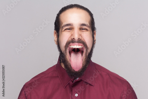 Portrait of funny crazy handsome man with beard in red shirt looking at camera with big open mouth and tongue out and screaming or shouting. indoor studio shot, isolated on gray background.